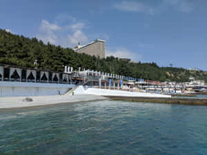 海岸度假勝地，有碧綠海水、白色卵石海灘和山腰酒店，在晴朗藍天下 — 海灘 壁紙