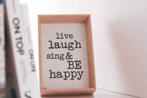 木框勵志語錄標牌，寫著 live laugh sing & BE happy，旁邊有書本。 — 3:2 壁紙