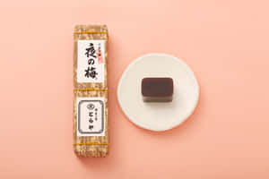 一個日本梅子醬包裝和一個深色糖果放在白色盤子上，背景是粉紅色。 — MacBook Air M3 壁紙