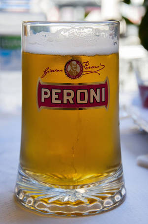 一個Peroni啤酒杯，裝有金黃色液體和白色泡沫，放在戶外桌子上。 — 啤酒 壁紙