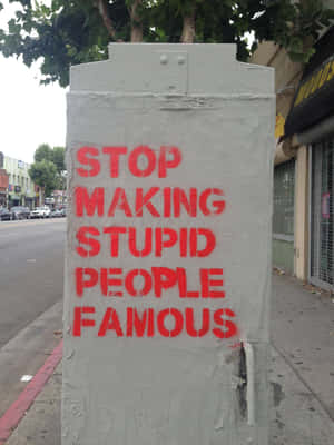 灰色公用箱上的紅色塗鴉寫著「STOP MAKING STUPID PEOPLE FAMOUS」，在城市人行道上。 — iPad 壁紙