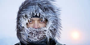 男人穿著結霜的毛皮兜帽，在雪白的冬日場景中凝視鏡頭。 — 寒冷 壁紙