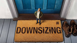 男人站在寫著 downsizing 的門墊上，附近有鞋子。 — 麥特·戴蒙 壁紙