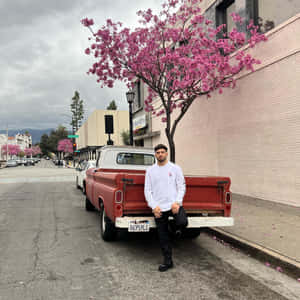 男人靠在復古紅色卡車旁，旁邊是粉紅色花樹，在城市街道上。 — 老福特卡車 壁紙