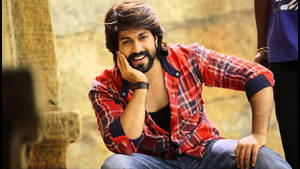 一個留鬍鬚的男人穿著紅色格紋襯衫，微笑著坐在戶外石頭附近。 — 寶萊塢 壁紙