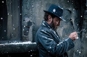 一個留鬍鬚的男人穿著外套和帽子，站在飄雪中，靠著灰色牆壁。 — 雪景 GIF 壁紙