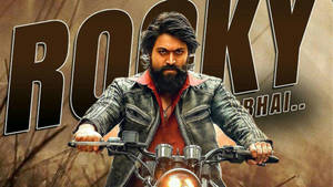 留鬍鬚的男人騎摩托車穿皮夾克，有 'Rocky Bhai..' 文字。 — The Krew 壁紙
