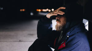 一個留鬍鬚的男人穿著連帽外套，夜晚獨自坐在街上，背後有模糊的燈光。 — 孤獨悲傷 壁紙