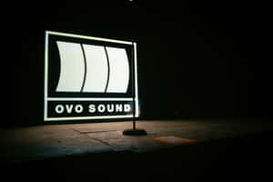 在黑暗的工作室環境中，一個明亮照明的 OVO SOUND 標誌。 — 3:2 壁紙