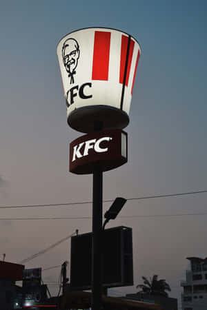 KFC桶形招牌在黃昏時分於城市環境中亮起，伴隨棕櫚樹和建築物。 — 榮耀MagicPad 3 Pro 壁紙