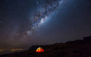 在星空下，一個發光的橙色帳篷位於山丘上，銀河清晰可見。 — 山脈 壁紙