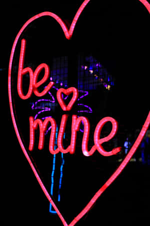 在黑暗的城市夜晚，一個明亮的粉紅色霓虹心形牌，上面寫著「be mine」。 — Vivo Pad 壁紙
