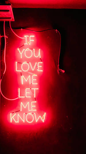 紅色霓虹燈牌，訊息為 'If you love me let me know'，置於深色牆上。 — 紅色美學 iPhone 壁紙