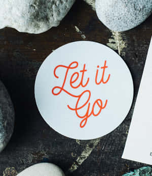 白色圓形貼紙，橙色草書文字寫著 Let it Go，被灰色石頭包圍。 — Galaxy Z Fold 壁紙