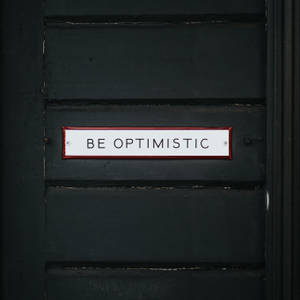 一個白色帶紅色邊框的標誌，上面寫著 BE OPTIMISTIC，位於深綠色木門上。 — 暗色 壁紙