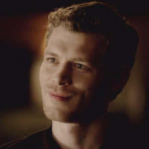 一個金髮、帶鬍渣的男人在昏暗溫暖的燈光下溫柔微笑的特寫。 — 噬血情人 壁紙