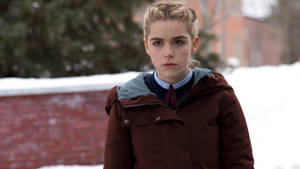 一個穿著冬季外套和校服的小女孩，站在磚牆附近的雪地中。
