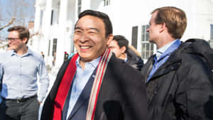 一個微笑的男人，戴著紅色圍巾和深色外套，與其他人在白色建築物附近戶外站立。 — 快樂 壁紙