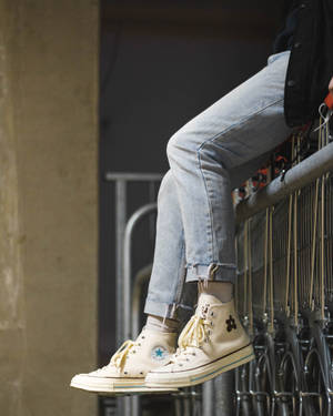 穿著奶油色運動鞋和牛仔褲的人坐在金屬欄杆上，附近有購物車。 — Converse 壁紙