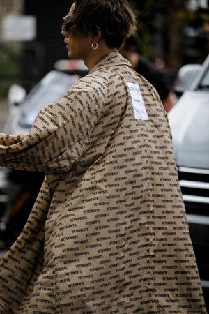 穿著帶有文字圖案的米色VETEMENTS外套在戶外行走的人。 — Vetements 壁紙