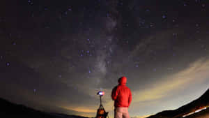 穿著紅色外套的人夜晚站在山上觀看星星和銀河。 — 美學天空 壁紙
