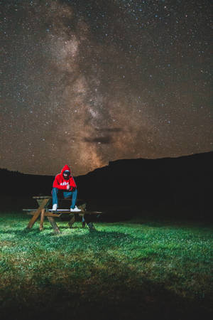 穿紅色連帽衫的人坐在野餐桌旁，星空下銀河拱橋 — 傷心 壁紙