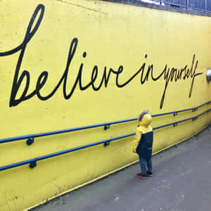 穿黃色外套的小孩看著黃色牆上的「believe in yourself」壁畫。 — 正向 壁紙