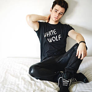 年輕男子穿著黑色「White Wolf」T恤和皮褲坐在白色床上。 — 大秀之王 壁紙