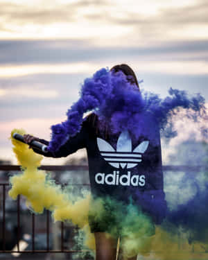 穿著黑色Adidas連帽衫的人在暮色中的屋頂上釋放紫色和黃色煙霧。 — 愛迪達 GIF 壁紙
