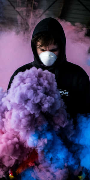 在工業空間中，穿著黑色連帽衫和白色口罩的人置身於彩色煙霧中。 — 瓦斯 壁紙