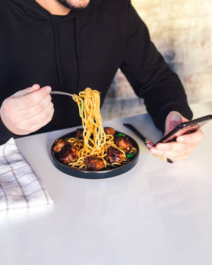 一個人在桌子旁使用智慧型手機吃帶有肉丸的麵條。 — 4:5 壁紙