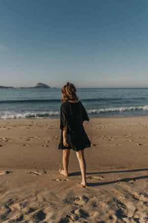 穿著黑色連衣裙的人赤腳走在沙灘上，面向夕陽下的海洋。 — 海灘度假 壁紙
