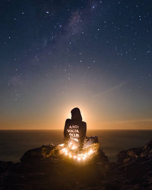 穿連帽衫的人夜晚坐在懸崖上，有串燈，觀看海洋上空的星星 — 反社會社交俱樂部 壁紙