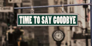 一個綠色路牌寫著「TIME TO SAY GOODBYE」，懸掛在城市街道上方，背景有一個時鐘。 — 再見 壁紙