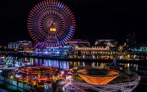夜間遊樂園，閃爍的遊樂設施和摩天輪倒映在水中。 — 城市燈光 壁紙