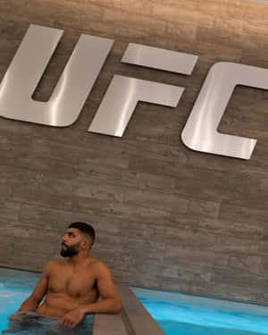一個留鬍子的男人在木牆上一個大型UFC標誌下的藍色水池中放鬆。 — 運動 壁紙