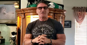 肌肉發達的男人戴著太陽眼鏡和 Ken Shamrock T恤，站在一個有獎盃的房間裡。 — 首頁 壁紙