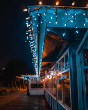 夜晚建築物下的木質步道，串燈發出藍色和溫暖的白色光芒。 — 4:5 壁紙