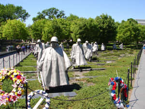 紀念公園中的灰色士兵雕像，配有色彩繽紛的花圈和綠樹，在藍天下。 — 陣亡將士紀念日 壁紙