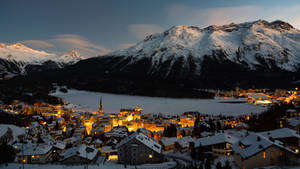 黃昏時被雪覆蓋的山村亮起燈光，附近有一個結冰的湖。 — Studio Display 壁紙