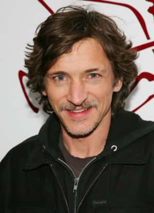 擁有波浪棕色頭髮和鬍鬚的男人穿著黑色外套微笑。 — 鷹眼 壁紙