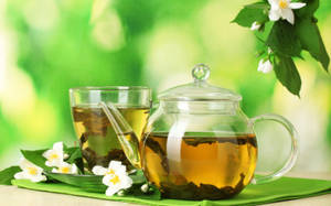玻璃茶壺和茉莉花茶杯，配上新鮮花朵和葉子，置於綠色布上。 — 花園 壁紙
