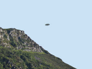 一個UFO在晴朗的藍天下飛越岩石密佈的綠色山脈。 — 太空船 壁紙