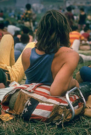 長髮的人坐在草地上，穿著藍色背心和美國國旗背包的戶外活動。 — Woodstock 壁紙