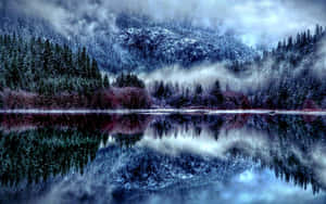 在霧雲下，平靜的冬日湖泊中倒映著積雪的山脈和松樹。 — 冬季風景 壁紙