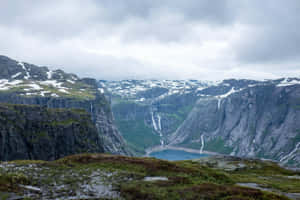 白雪覆蓋的山脈，瀑布在陰天天空下流入藍色湖泊。 — 山景 壁紙