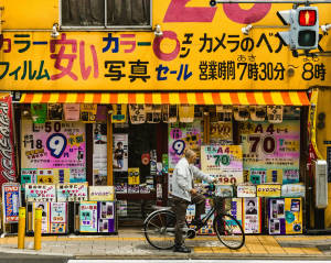 騎腳踏車的男人在色彩繽紛的日本相機店前停下來，店內有促銷標誌。 — 日本 壁紙