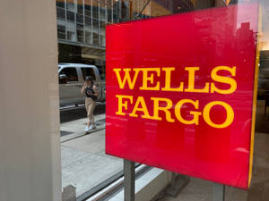 一個帶有黃色字樣的紅色 Wells Fargo 標誌反射出一個女人在城市人行道上行走。 — 街頭 壁紙