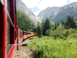 紅色和橙色的火車穿過一個有綠樹和岩石山峰的山谷。 — 科羅拉多 壁紙