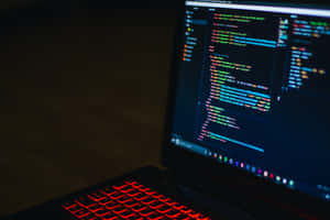 在黑暗房間中，帶有紅色背光鍵盤和發光代碼的筆記型電腦 — MacBook Pro M2 壁紙